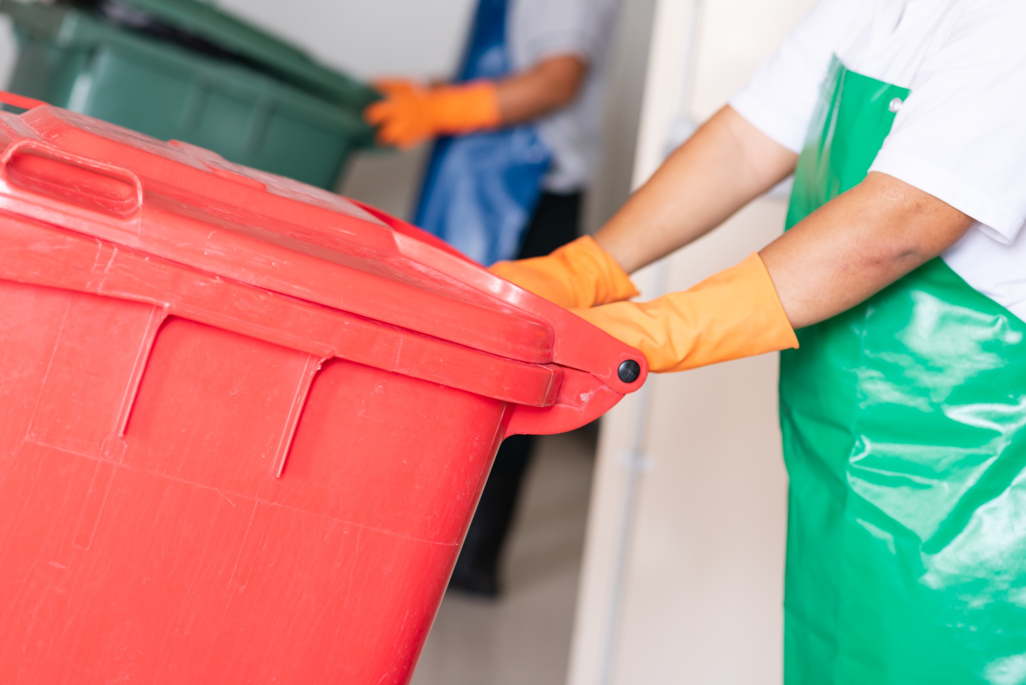 A worker in orange gloves lifts a red trash bin while another in a green apron assists in waste management, highlighting cleanliness and organization.